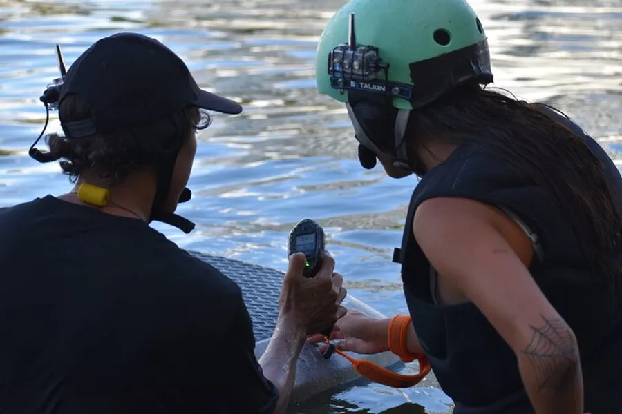 Two individuals, one wearing a helmet equipped with communication gear, are handling some equipment near a body of water.