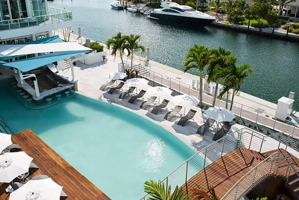 The image shows a luxurious outdoor pool area with loungers and palm trees overlooking a marina with a yacht