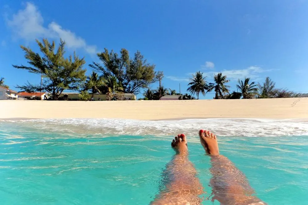 A person is relaxing in clear shallow waters at a sandy beach with their feet extended towards a blue sky and tropical backdrop