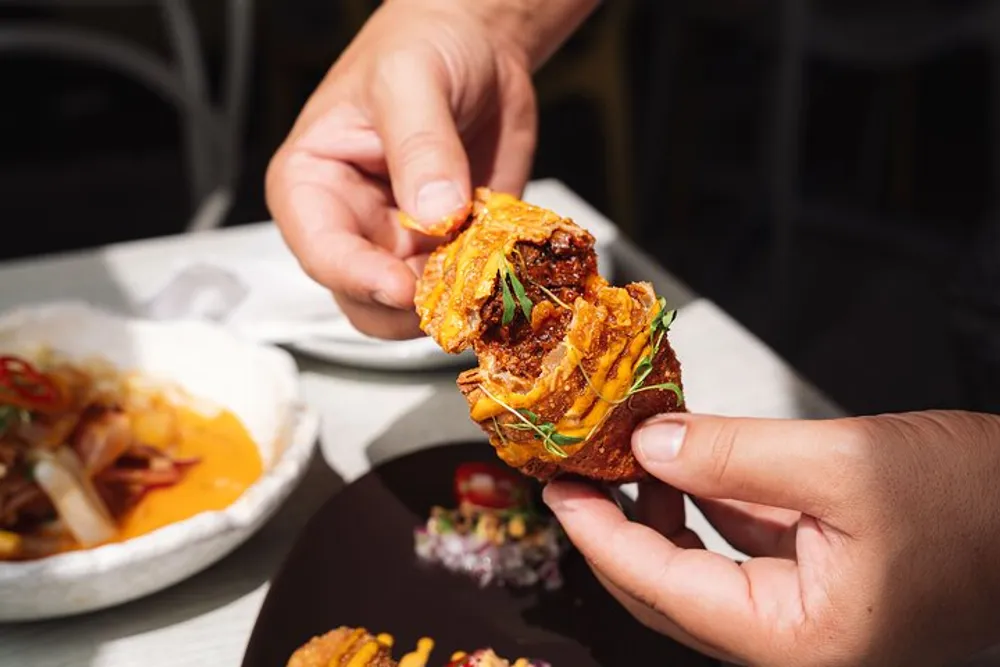 Someone is holding a savory stacked mini sandwich or appetizer between their fingers showing a close-up view of the food with garnishes and other dishes blurred in the background