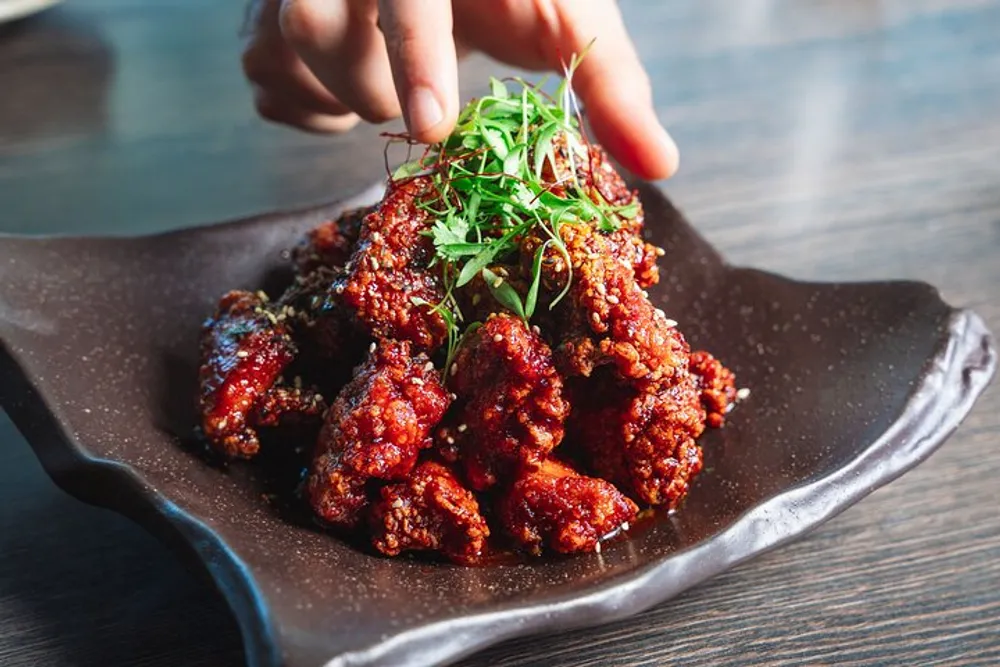 A person is garnishing a plate of crispy glazed chicken with fresh herbs
