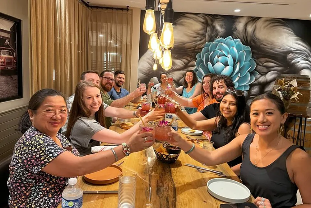 A group of smiling people are toasting with drinks around a dining table in a warmly lit room with a large floral mural on the wall