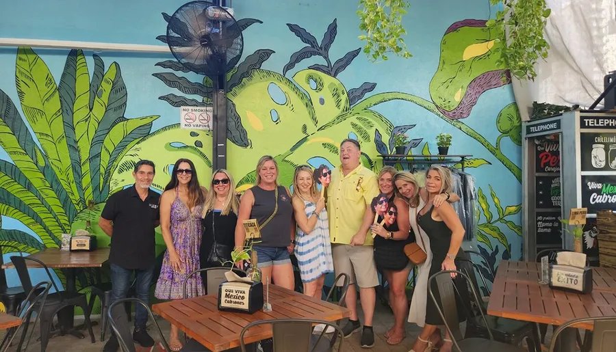 A happy group of people are posing for a photo at an outdoor venue with a colorful mural of a green dinosaur in the background.