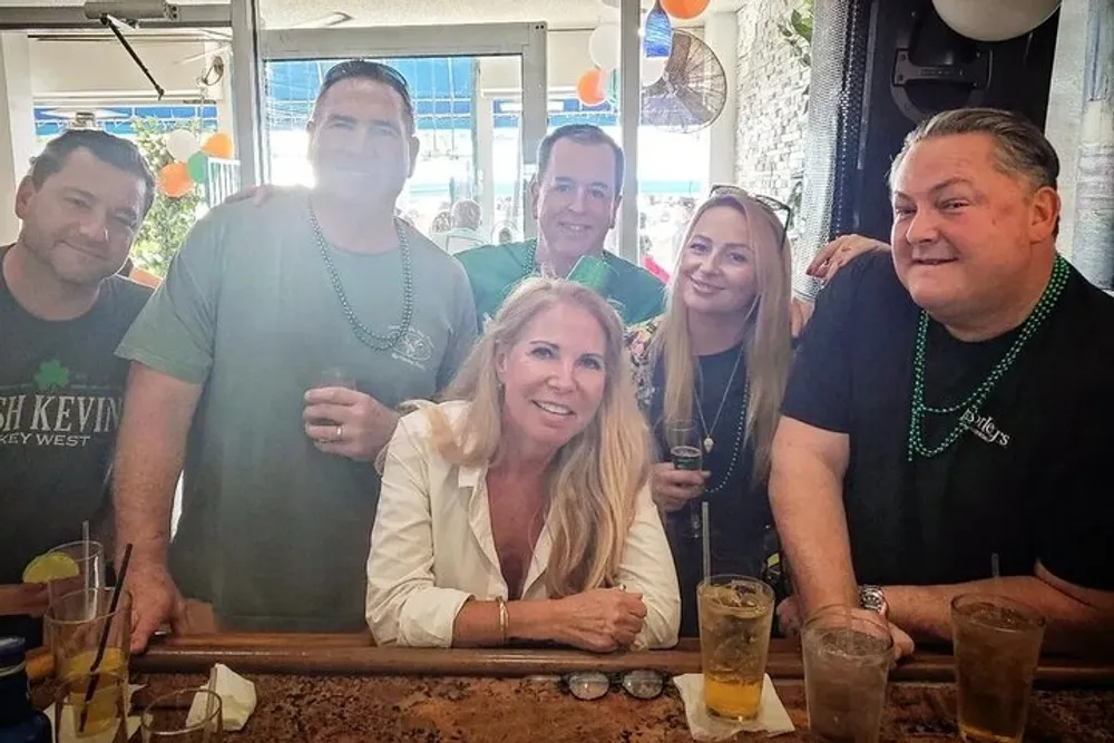 A group of smiling people are enjoying drinks together at a bar with some wearing green beads suggesting a festive or celebratory occasion