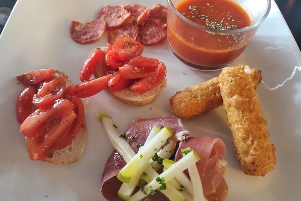 The image shows a variety of appetizers on a white plate including sliced tomatoes on bread cured meats cheese sticks pepperoni slices and a cup of sauce