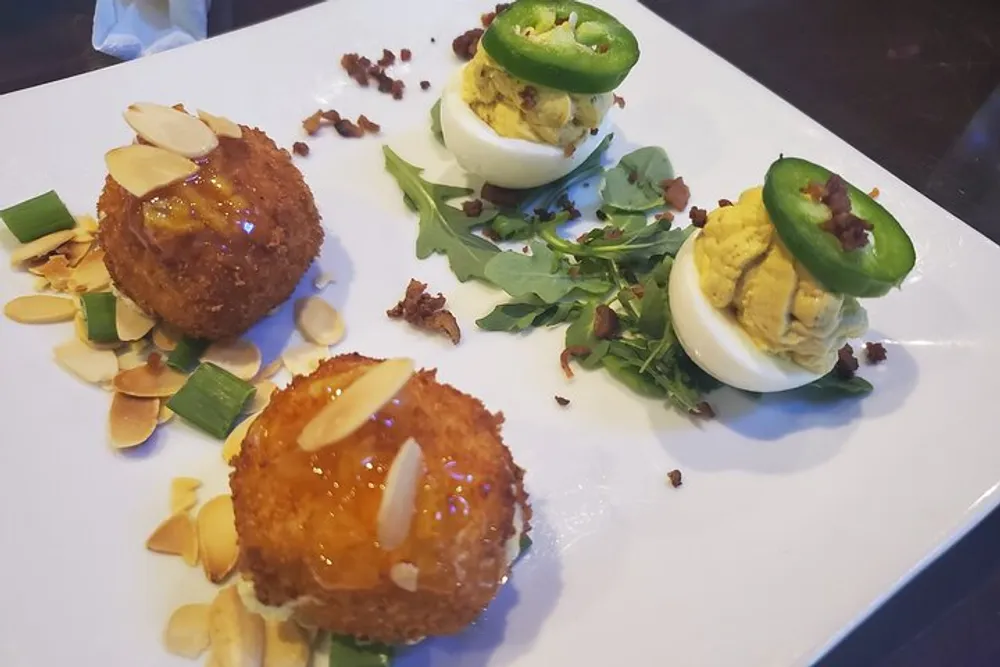 The image features a plate of appetizers with two golden-brown almond-topped deep-fried balls and a pair of deviled eggs garnished with green jalapeo slices all displayed on a bed of greens