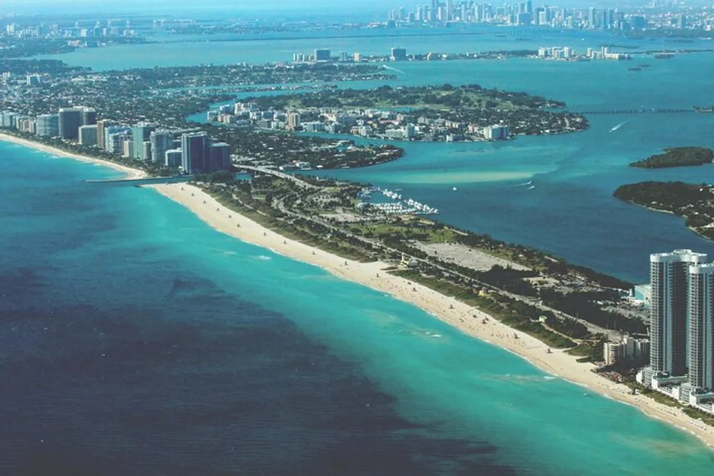 The image shows an aerial view of a coastal cityscape with numerous buildings adjacent to a long stretch of beach along a vivid blue ocean and inlet possibly taken from an airplane