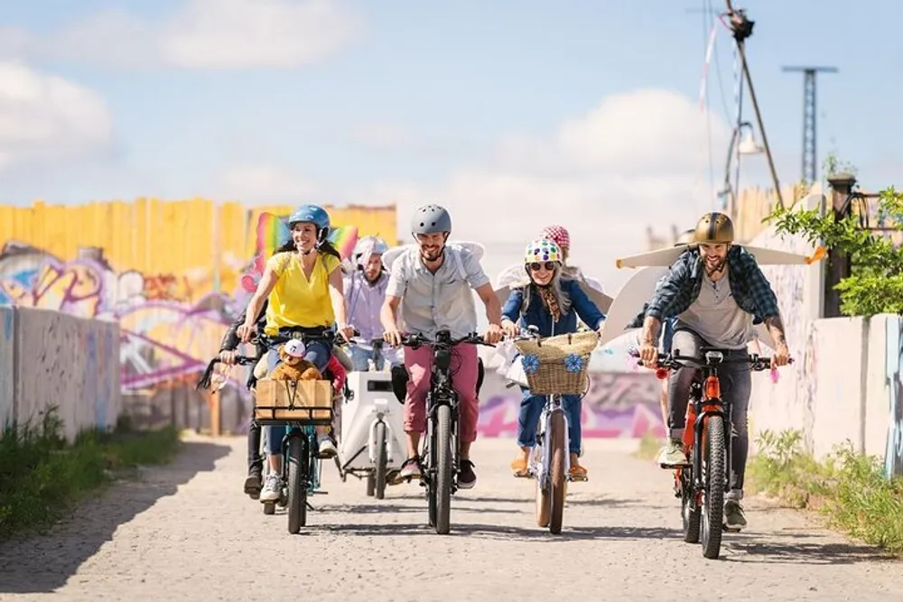 A group of people are cheerfully riding bicycles down a path with colorful graffiti art on the walls in the background