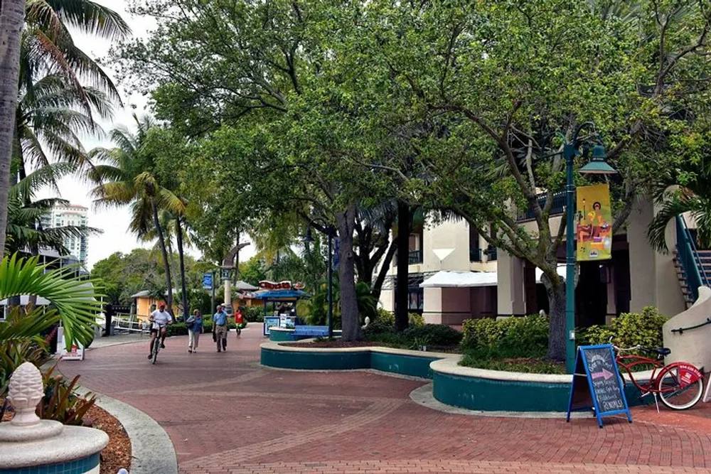 The image shows a pedestrian walkway lined with trees with a few people walking a bicycle parked on the side and buildings in the background under an overcast sky