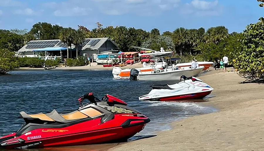 The image shows a sunny beach scene with personal watercrafts and boats near the shore and a building with solar panels in the background.