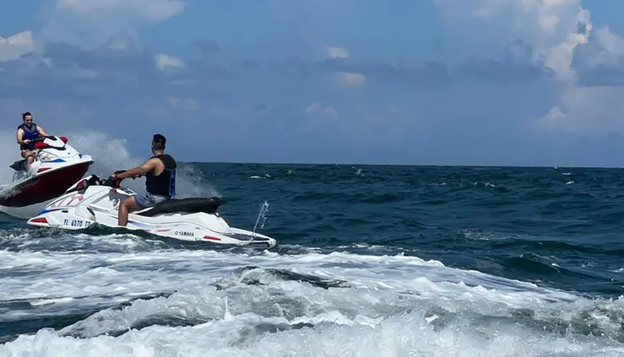Two people are riding jet skis on choppy sea waters under a partly cloudy sky.