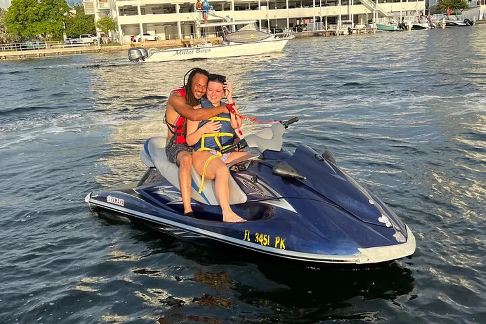 Two people are smiling and embracing on a jet ski with waterfront buildings in the background during late afternoon sunlight