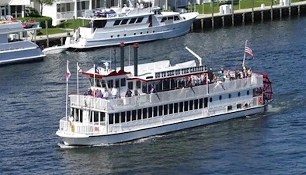 A paddlewheel riverboat filled with passengers is cruising on a river past docked yachts and waterfront properties
