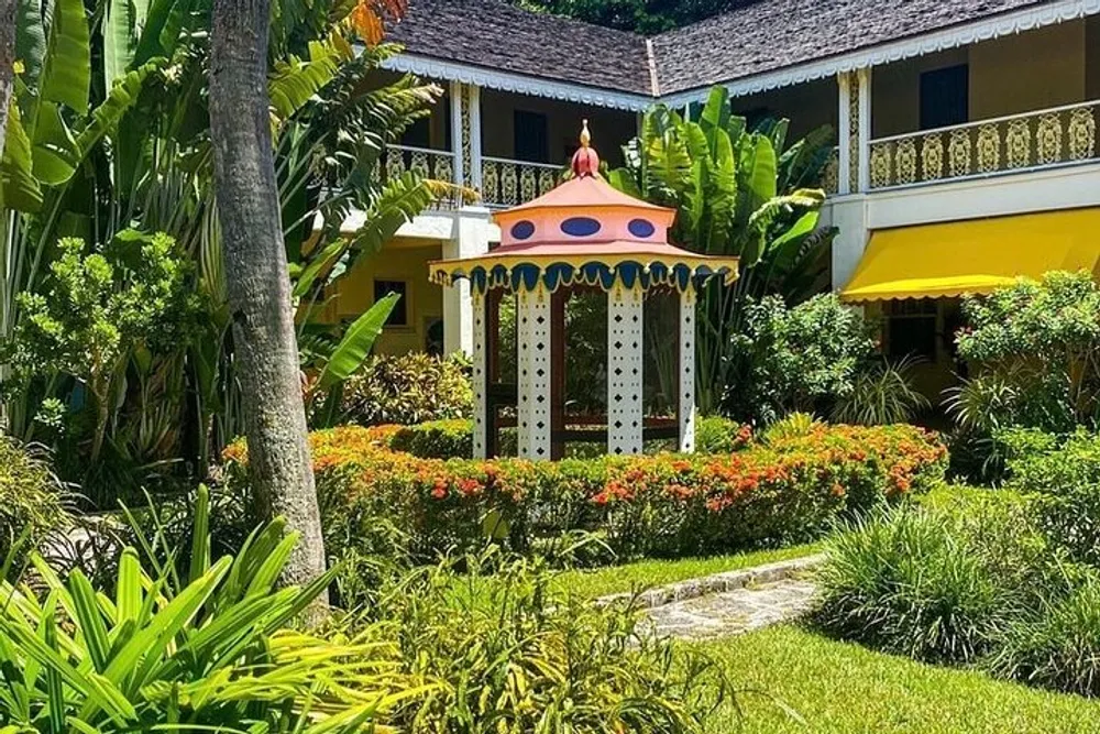 A colorful garden gazebo sits in front of a vibrant yellow building surrounded by lush tropical vegetation