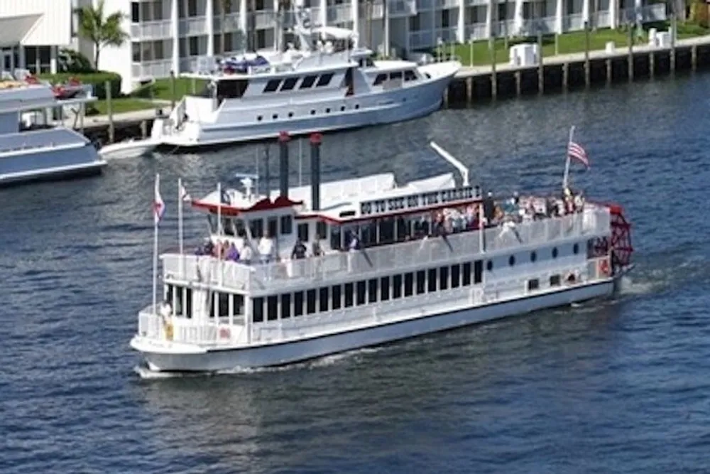A paddle wheel riverboat filled with passengers is cruising along the water with other boats and waterfront buildings in the background