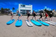 A group of people is engaging in a paddleboard yoga pose on the beach, with clear blue skies above them and a modern building in the background.