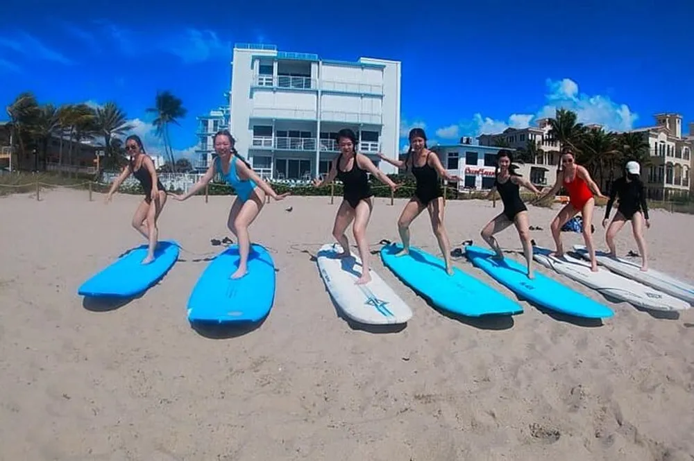 A group of people is engaging in a paddleboard yoga pose on the beach with clear blue skies above them and a modern building in the background