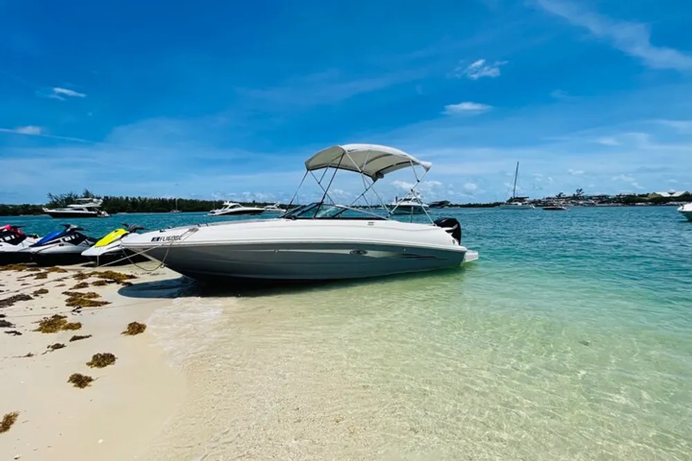 A boat and a jet ski are beached on a sandy shore with clear blue waters and other boats in the background under a partly cloudy sky