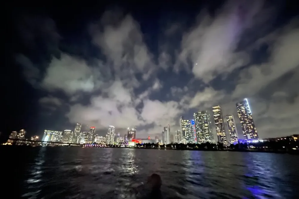 The image shows a nighttime city skyline viewed across a body of water with a dramatic cloudy sky overhead
