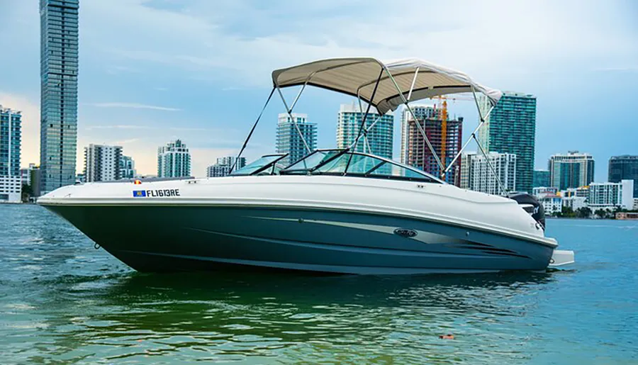 A white motorboat with a bimini top is moored on calm waters with a backdrop of a modern city skyline.