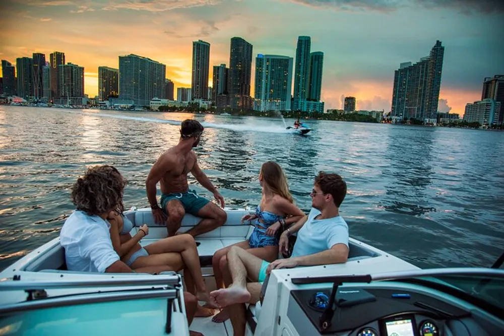 A group of people is enjoying a boat ride with a view of a spectacular sunset over a city skyline while another person is jet skiing nearby