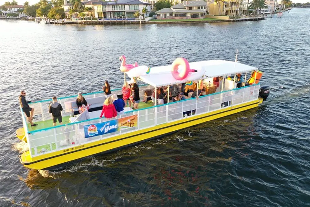 A group of people enjoys a sunny boat ride on a colorful watercraft adorned with a flamingo float cruising along a waterway flanked by residential buildings
