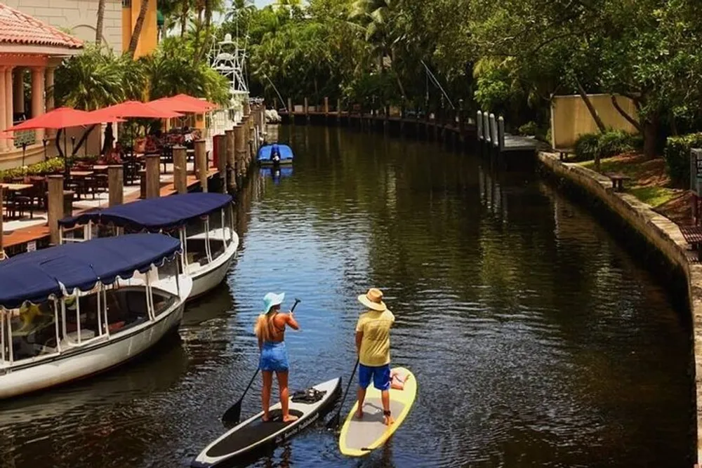 Two people are paddleboarding along a calm waterway flanked by moored boats and tropical vegetation