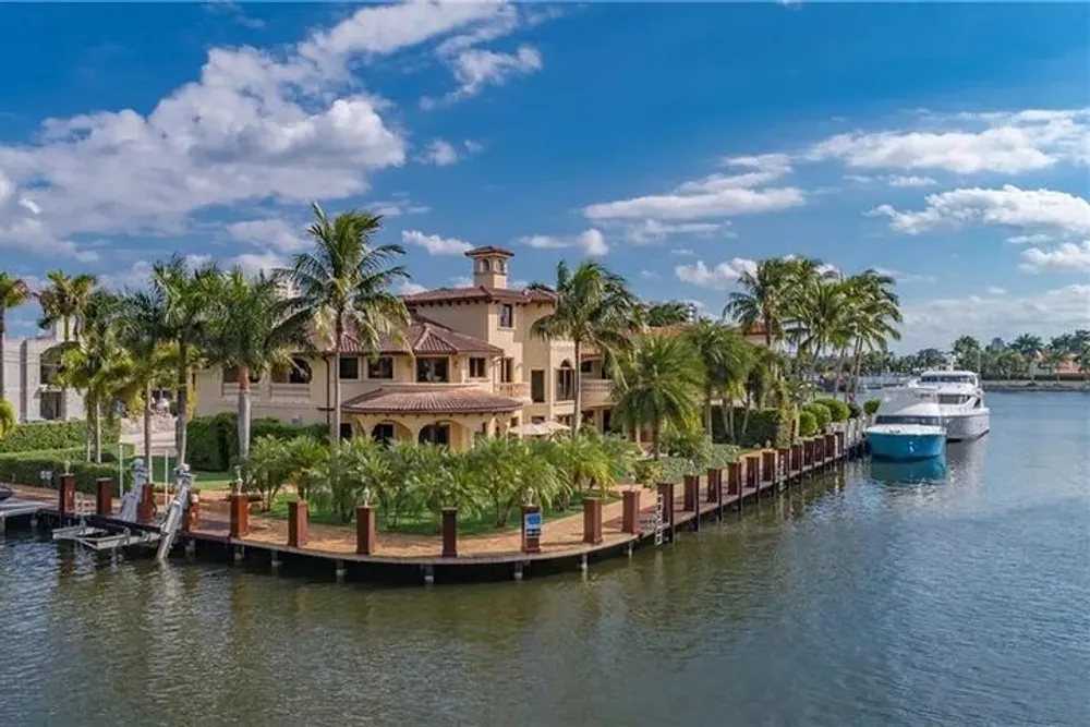 A luxurious Mediterranean-style villa is nestled among palm trees on a waterfront property with a large yacht docked nearby