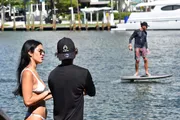 A person in a bikini is engaged in a conversation with an individual wearing a black shirt and hat, while another person is paddleboarding in the background near docked boats.