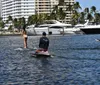 A person in a bikini is engaged in a conversation with an individual wearing a black shirt and hat while another person is paddleboarding in the background near docked boats