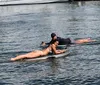 A person in a bikini is engaged in a conversation with an individual wearing a black shirt and hat while another person is paddleboarding in the background near docked boats