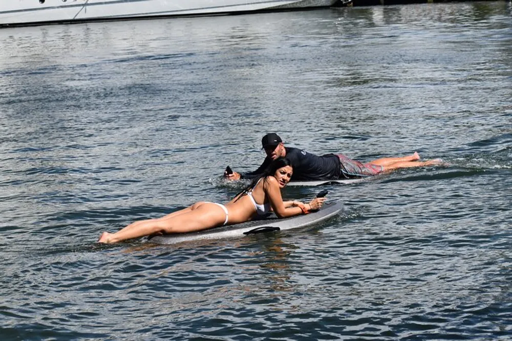 A person is lying on a surfboard while another person holds onto it both in the water near a dock