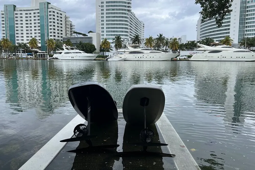 The image shows the stern of a boat with its propellers and rudder exposed docked in a marina with modern buildings and luxury yachts in the background under an overcast sky