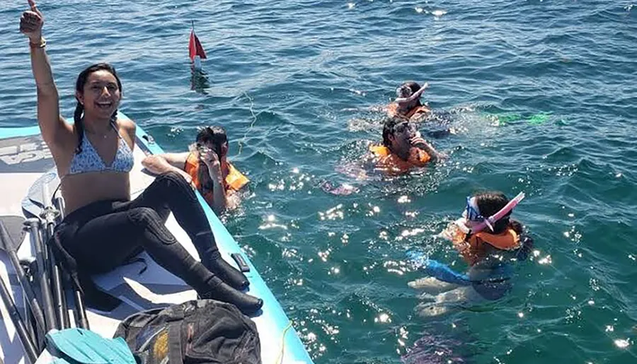 A group of people are enjoying water activities, with some wearing snorkeling gear in the sea while a person seated on the edge of a boat is raising her hand in a gesture of excitement or victory.