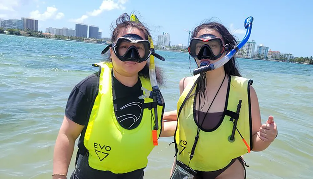 Two individuals are standing in shallow water wearing snorkeling gear and yellow life jackets with a coastal cityscape in the background