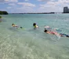 The image shows a group of people snorkeling in clear shallow waters near a tropical coastline with buildings in the background