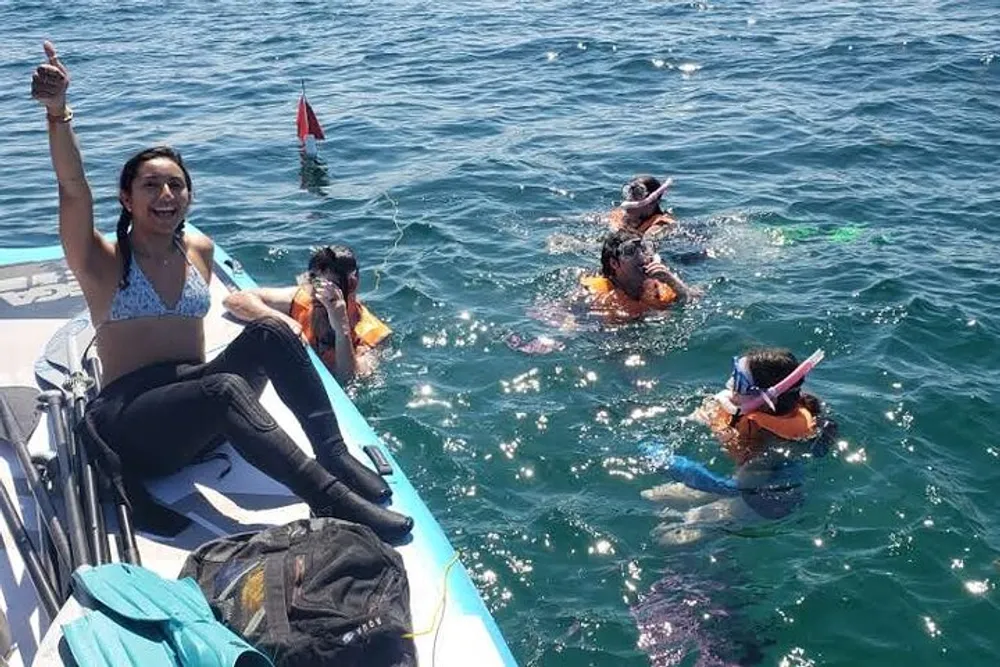 A group of people enjoy snorkeling in clear blue waters near a boat with one person sitting on the edge of the boat raising her hand in a cheerful gesture