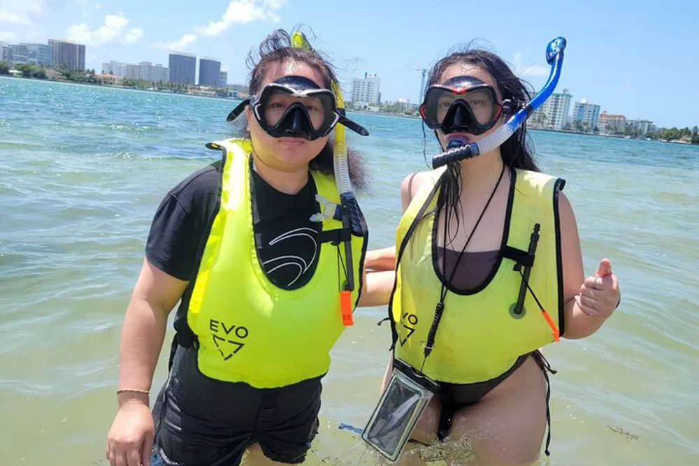 Two individuals are standing in shallow water wearing snorkeling gear and bright yellow life vests possibly getting ready for or returning from a snorkeling adventure