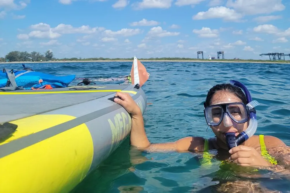 A person equipped with a snorkeling mask and a snorkel is smiling at the camera while holding onto a kayak in clear blue waters