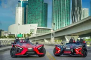 Two Polaris Slingshot three-wheeled vehicles are parked on an urban road with skyscrapers and a bridge in the background.