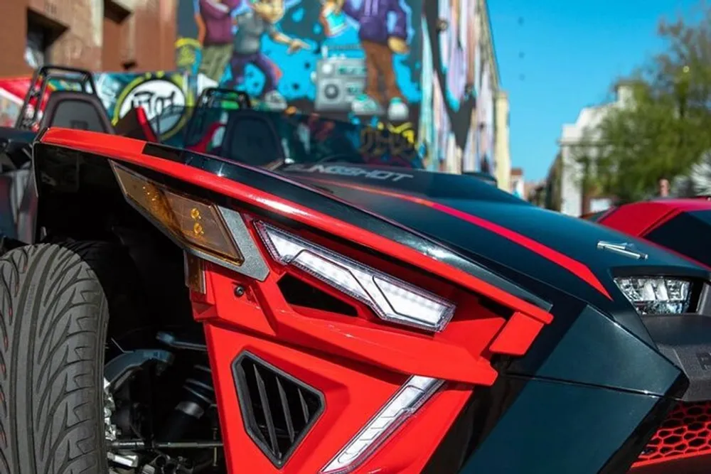 A close-up of a red and black sporty vehicle with futuristic headlight design parked on a street with vibrant graffiti art in the background