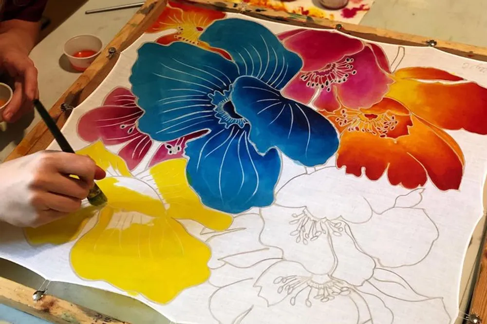 A persons hand is seen painting a vibrant watercolor of large colorful flowers on paper which is stretched and secured on a wooden frame