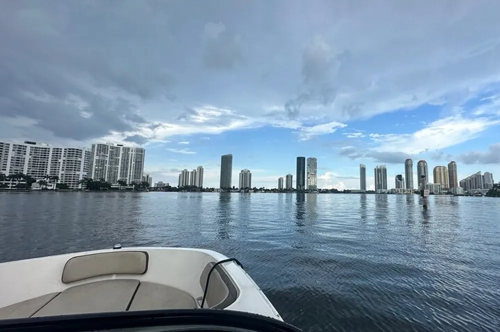 A view from the bow of a boat approaching a city skyline across a calm body of water with a partly cloudy sky above