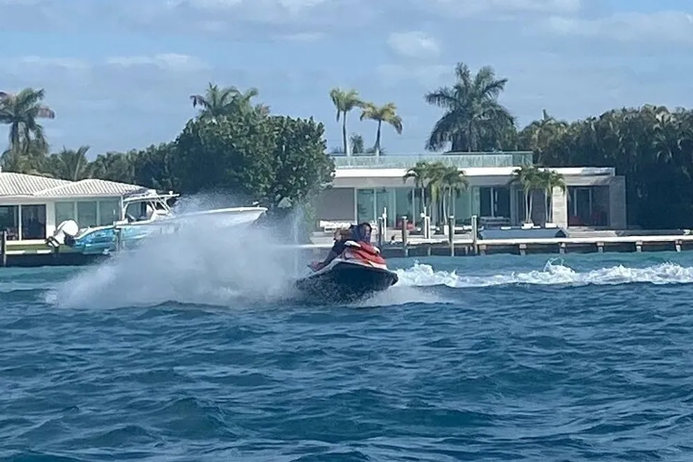A person on a jet ski is making a sharp turn creating a large spray of water with a backdrop of a coastal residence