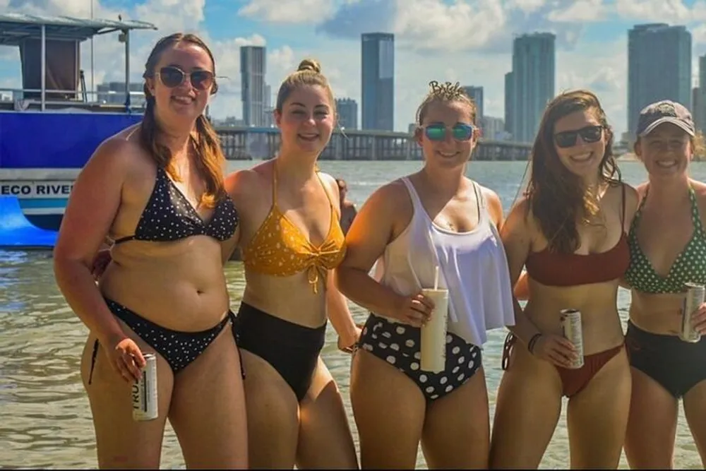 A group of five women in swimwear are smiling for a photo by the water with a city skyline in the background