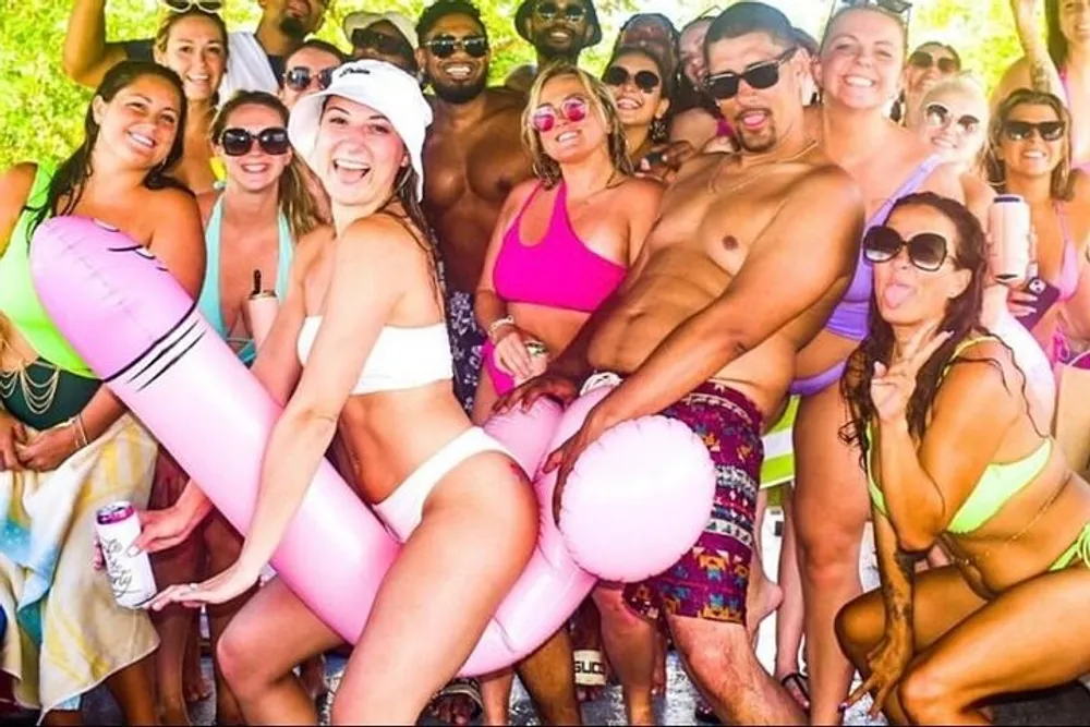 A group of cheerful people in swimwear is posing for a photo at a beach or pool party with some holding drinks and inflatable toys