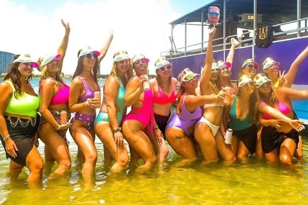 A group of people in swimwear and matching sun visors are joyfully posing in shallow water with a boat in the background giving the impression of a festive beach outing