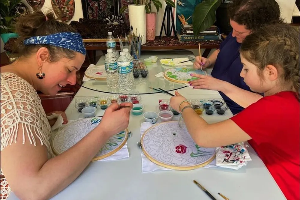Three people are painting on embroidery hoops at a crafting table with various paint jars and brushes around them