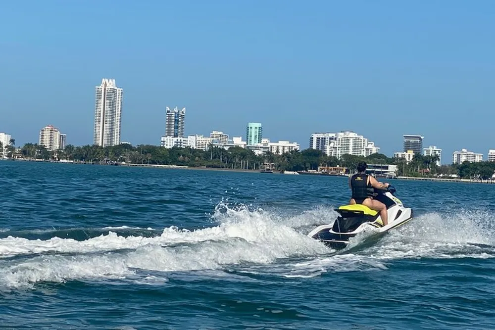 A person is riding a jet ski on the water with a coastline and buildings in the background