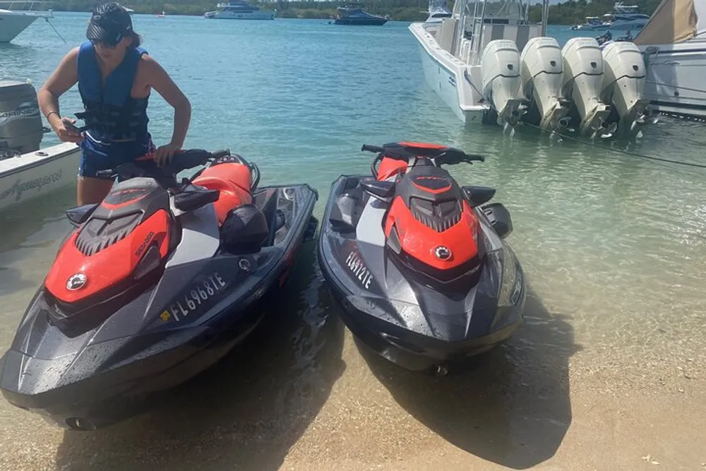 A person stands by two jet skis on a sunny beach with boats moored in the background